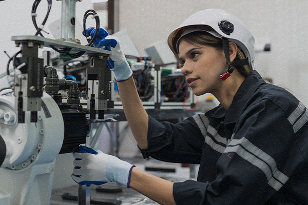 Woman in hard hat and safety gear uses gloved hands to examine a piece of machinery.