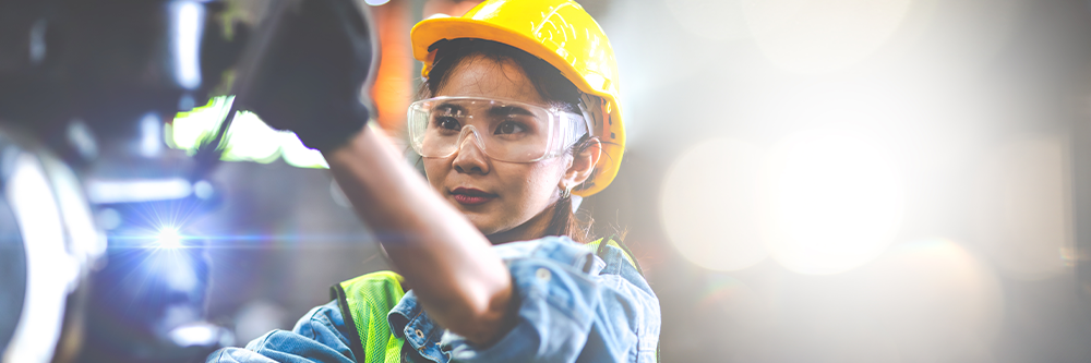 Woman in hard hat and gloves holds a metal tool to repair a piece of machinery.