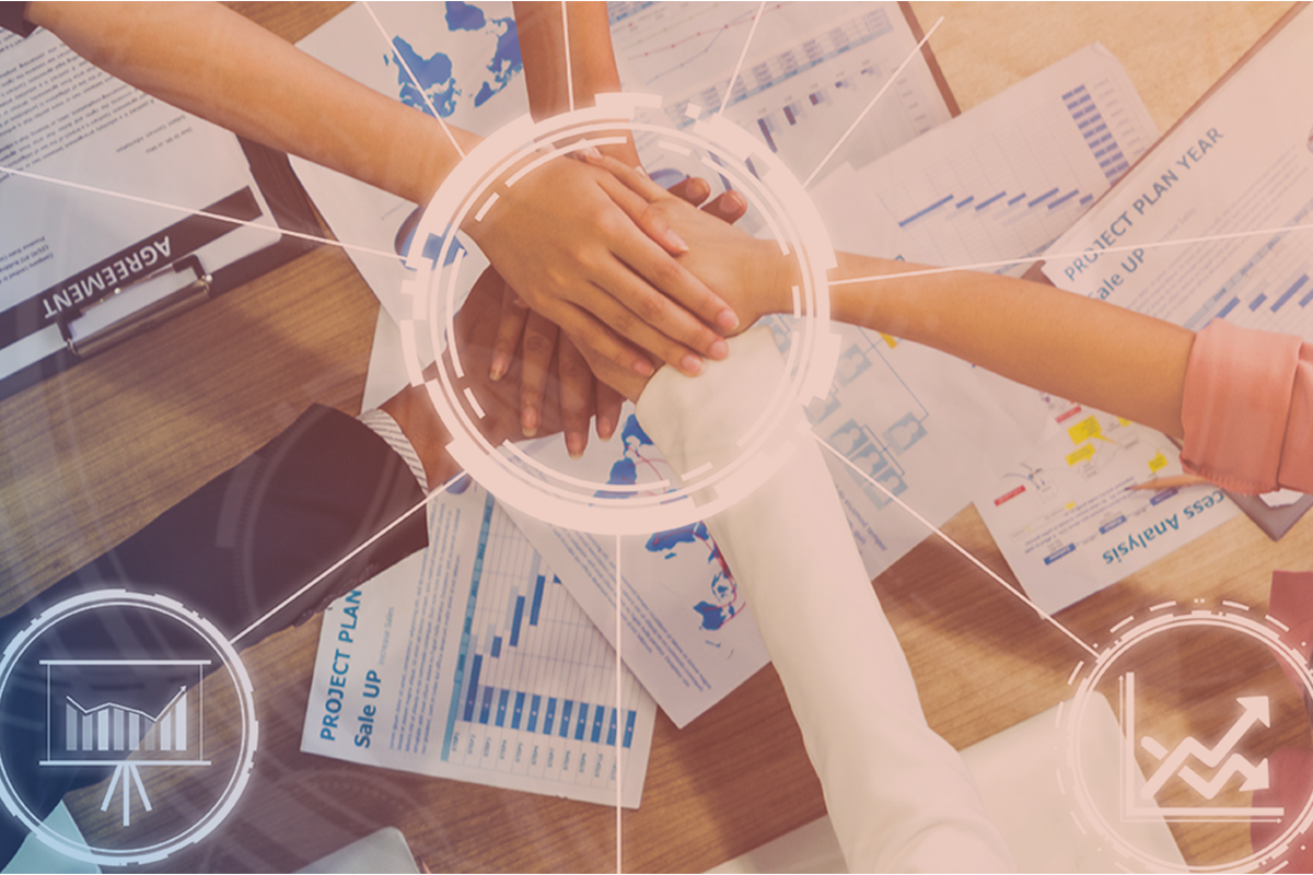 Hands join in the center of a table covered in business papers showing graphs, planning, maps, and a business agreement. Manufacture Nevada logo is overlaid in the corner.