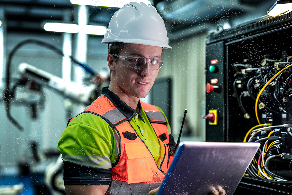 A man in hard hat, glasses, and safety vest holds a laptop near a control panel with wires.