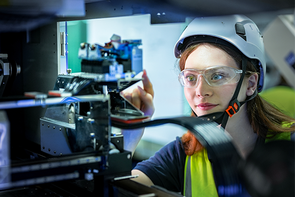 Woman in safety glasses and hard hat points at the inner mechanisms of a machine.