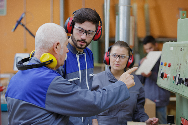 Man in jumpsuit and work headphones gestures towards a machine. A man and woman dressed in work clothes observe him.