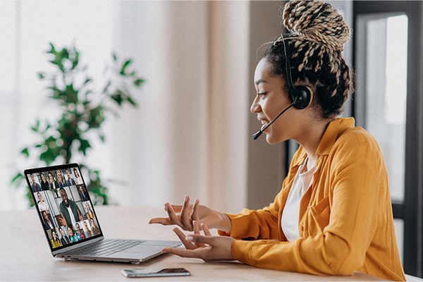 Woman at table wears a headset and watches a group Zoom meeting on a laptop.