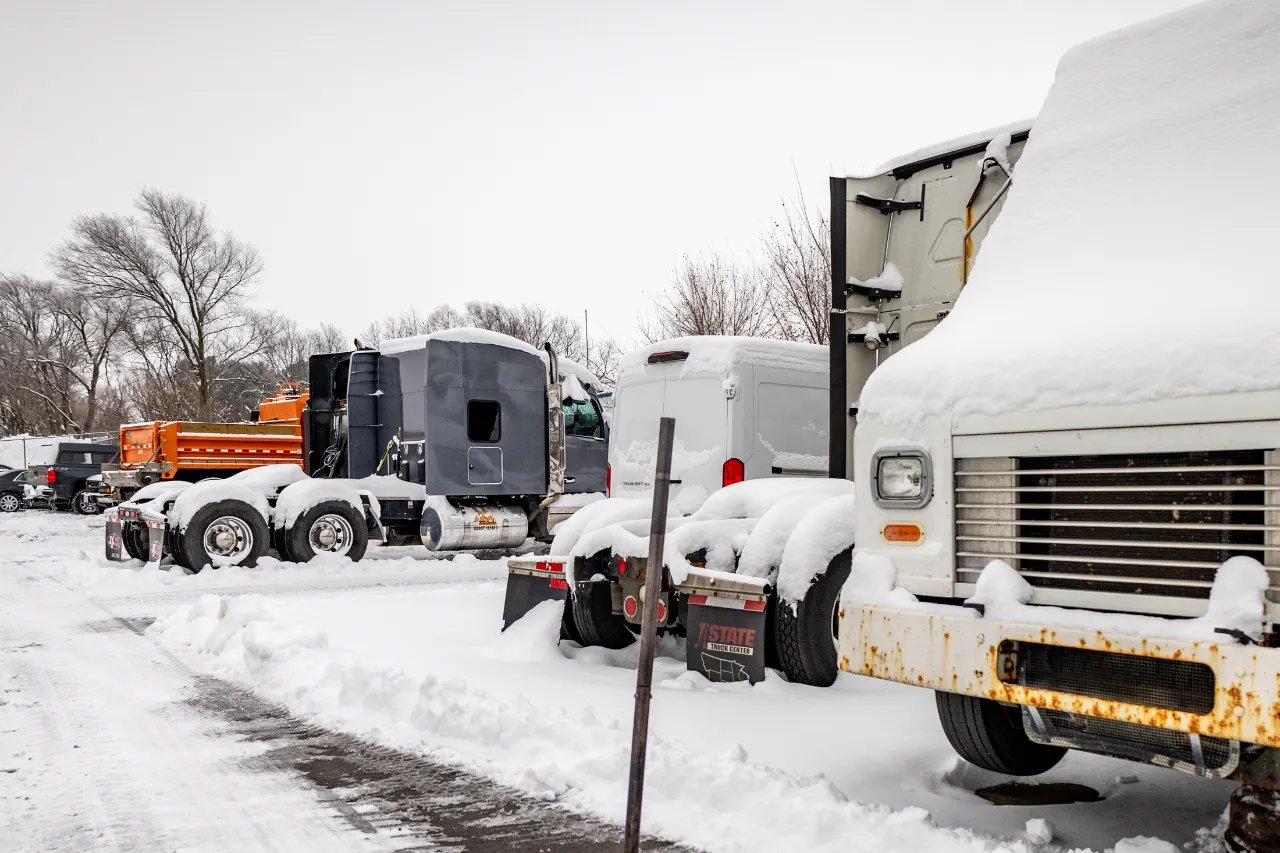 Snow-covered heavy-duty trucks and trailers parked in a New Hampshire lot awaiting winter maintenance.