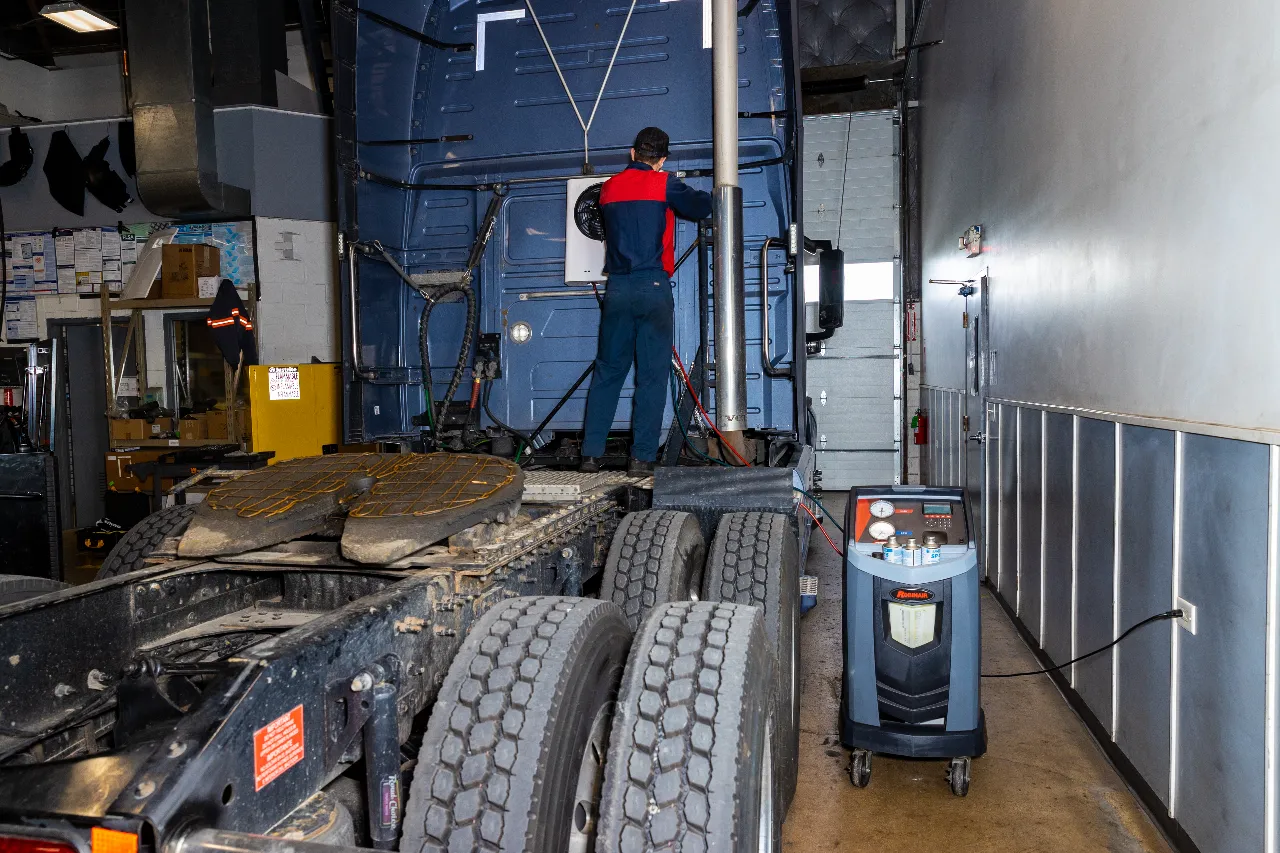 Heavy-duty A/C service, technician connecting recovery machine to semi-truck cab inside repair shop.