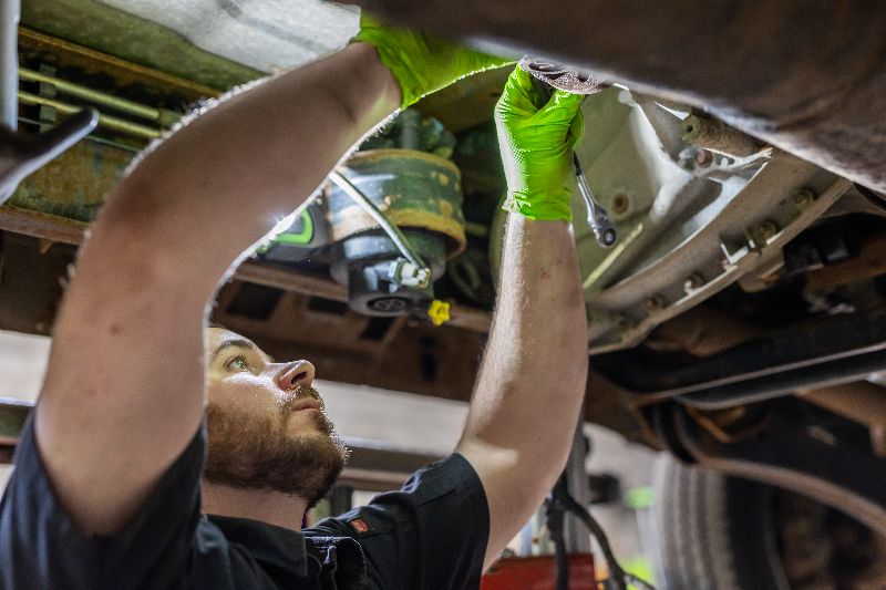 Diesel technician wearing gloves working underneath truck, using tools to service drivetrain components during maintenance in professional repair shop setting