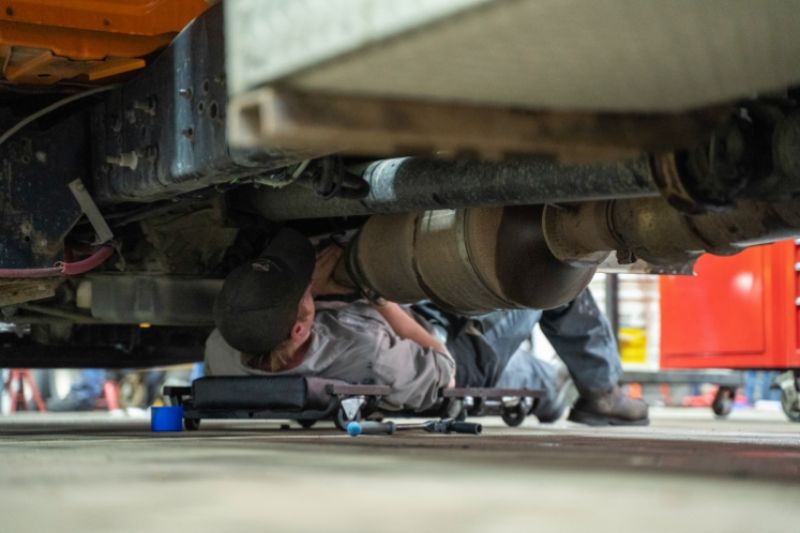 Technician working beneath a heavy-duty truck in a repair shop, inspecting and servicing exhaust and emissions system components.