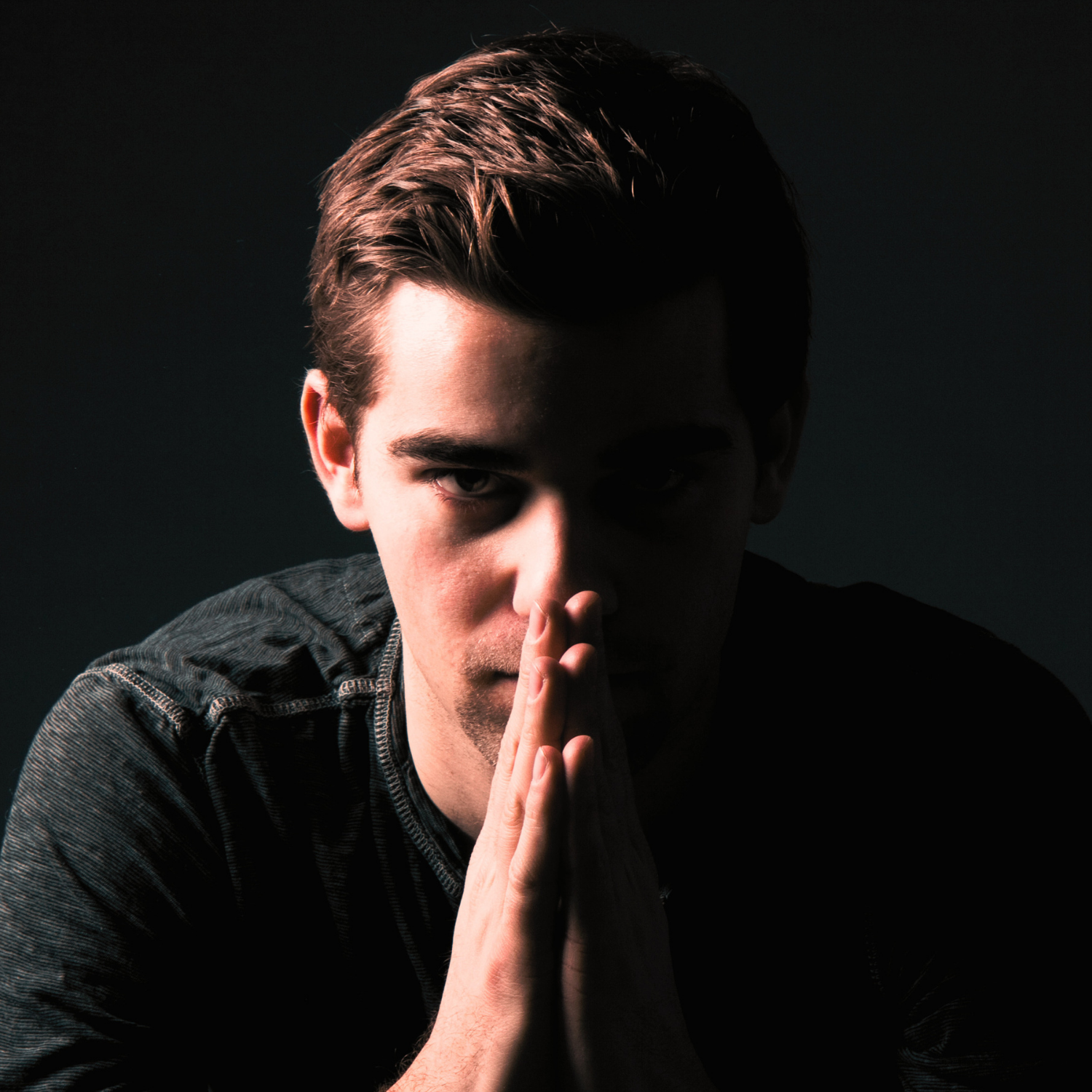 Young man in dark shirt with hands pressed together in front of face, low lighting on one side.
