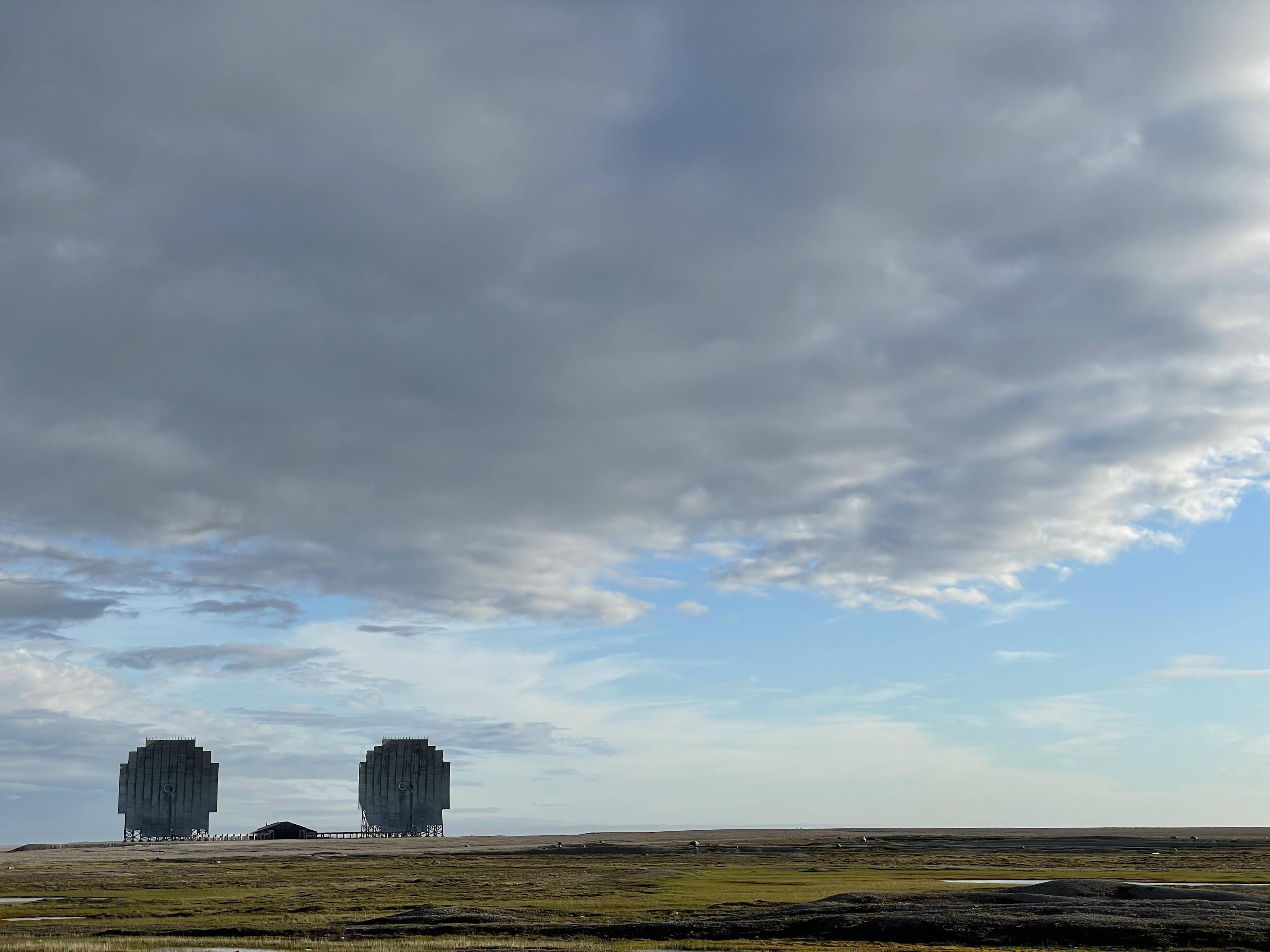 Two large, circular radar dishes on a flat grassy plain under a cloudy sky with patches of blue.