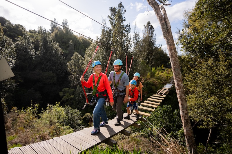family walking on a swing bridge attached to line through native forest in Okere Falls, Rotorua