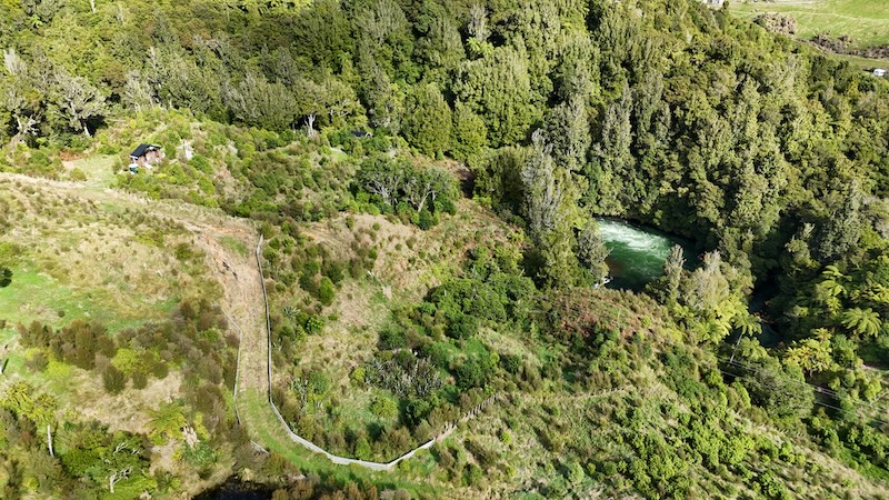 Aerial view of the Rotorua Ziplines course at Ōkere Falls, showing twin ziplines threading through a mosaic of native bush and open hillside. The Kaituna River is visible on the left, and a zipline platform shelter sits on the ridge above. The landscape shows active regeneration, with native ferns, flax, and young trees established across the slope.