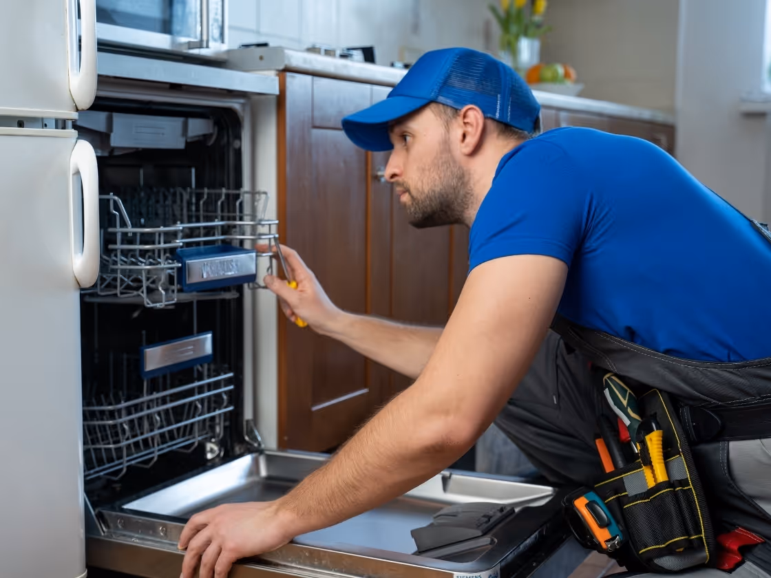 Technician in blue repairing dishwasher with tool belt in kitchen