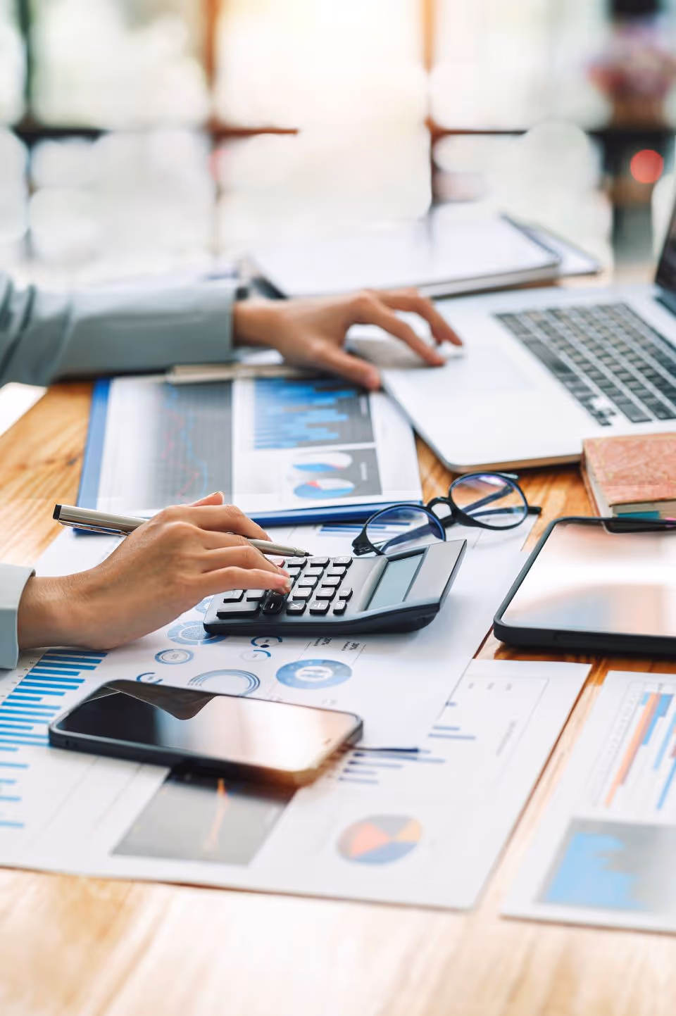 Person using calculator and laptop on a desk with printed charts, smartphone, tablet, and eyeglasses.