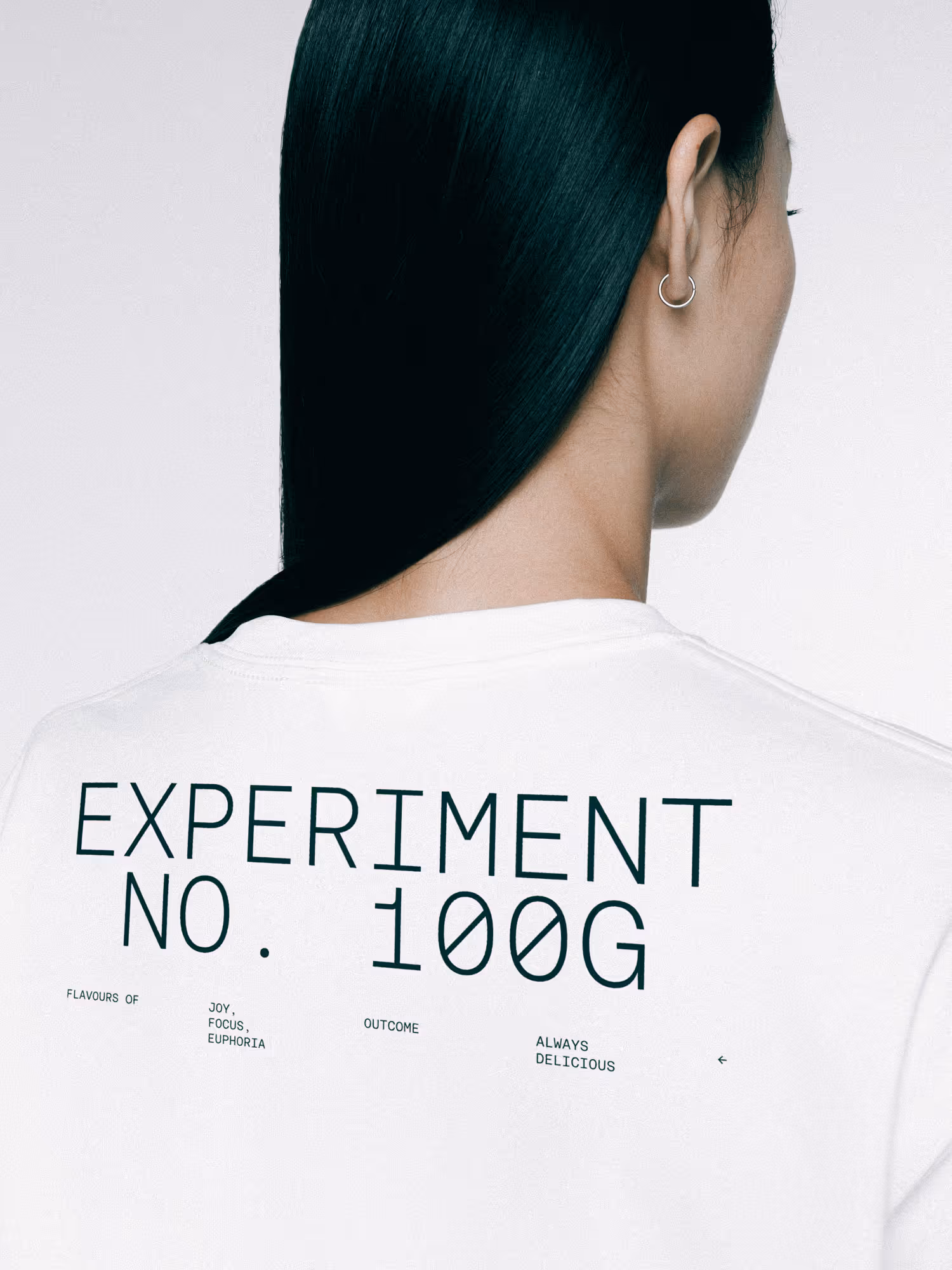 Back view of person with long black hair wearing a white shirt with black text reading 'EXPERIMENT NO. 100G' and smaller text including 'FLAVOURS OF JOY, FOCUS, EUPHORIA', 'OUTCOME', and 'ALWAYS DELICIOUS'.