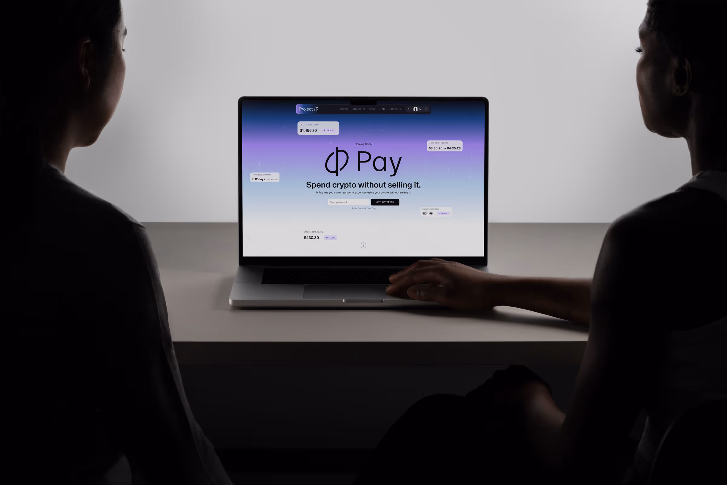Two people looking at a laptop displaying a crypto payment app with the text ‘Spend crypto without selling it.’