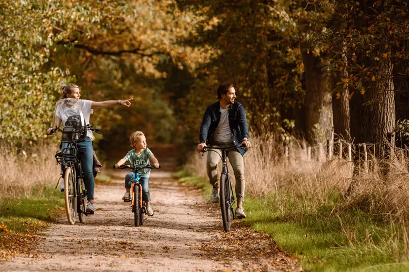 Gezin met moeder, vader en kind fietst op een bosrijk pad in de herfst, moeder wijst naar links.