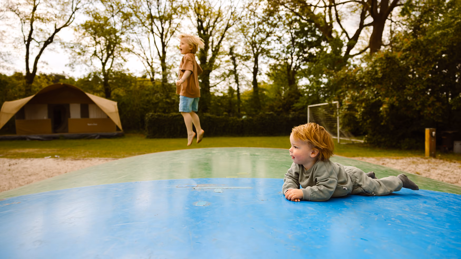Twee kinderen spelen op een luchtkussen buiten, met tent en voetbalgoal op de achtergrond.
