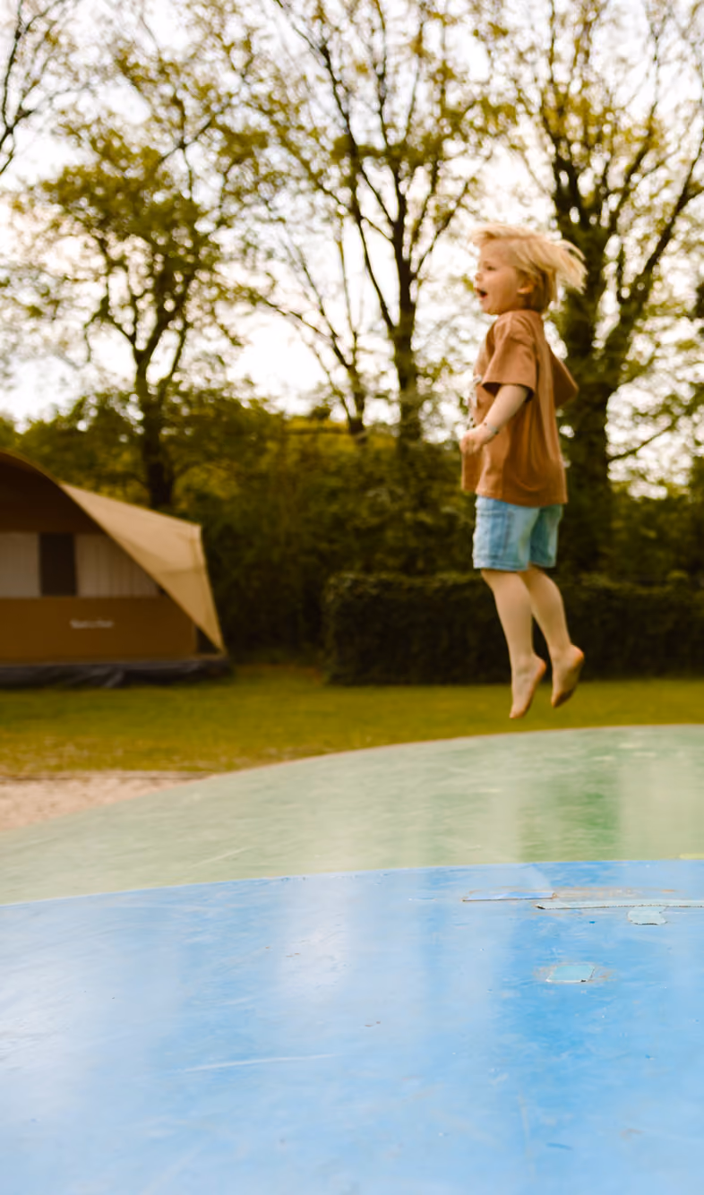 Twee kinderen spelen op een luchtkussen buiten, met tent en voetbalgoal op de achtergrond.