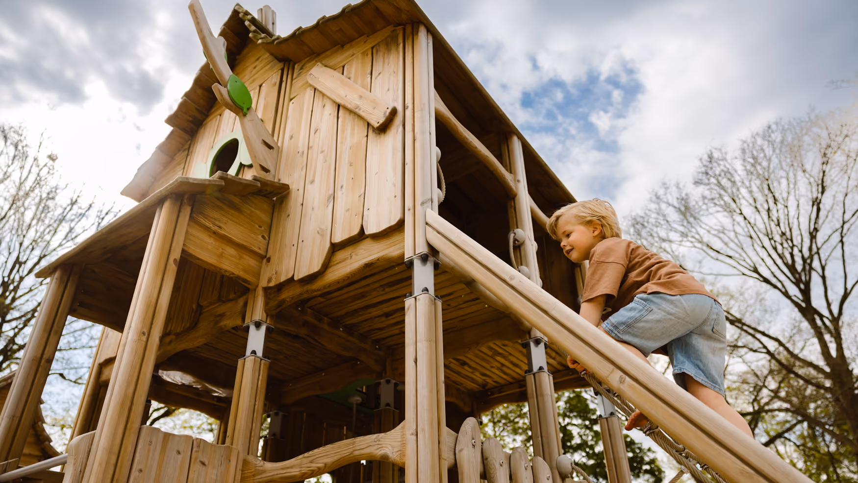 Kind klimt op houten speelhuis buiten onder bewolkte lucht met bomen.