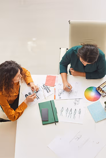Two women collaborating on fashion sketches at a white table with a color wheel and notebooks.