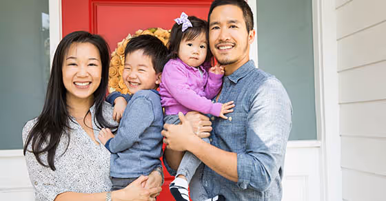 Smiling family of four standing in front of a house entrance with a red door, parents holding their young children