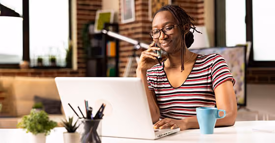 woman sitting at table with laptop and coffee cup talking on cell phone