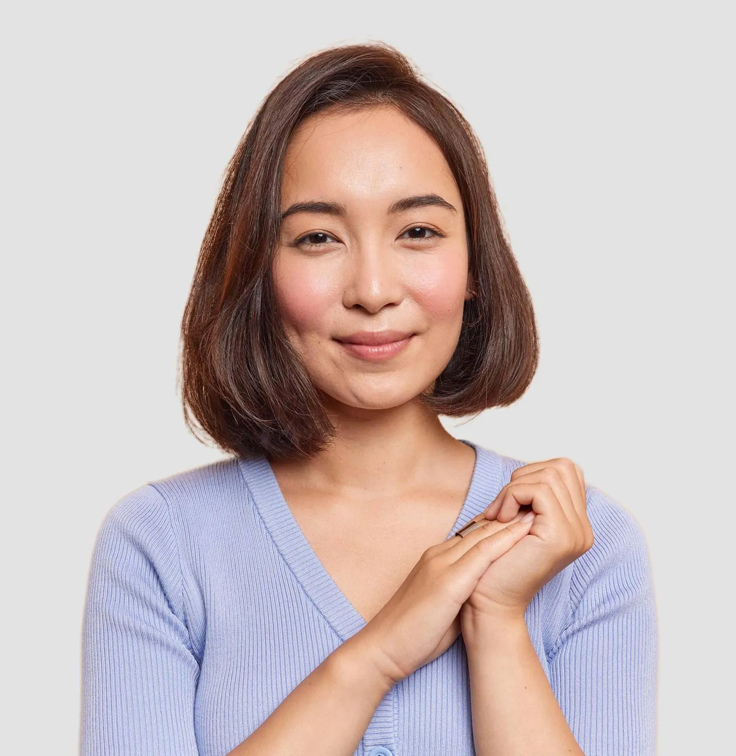 Smiling woman with shoulder-length brown hair wearing a light purple V-neck sweater, gently clasping her hands.