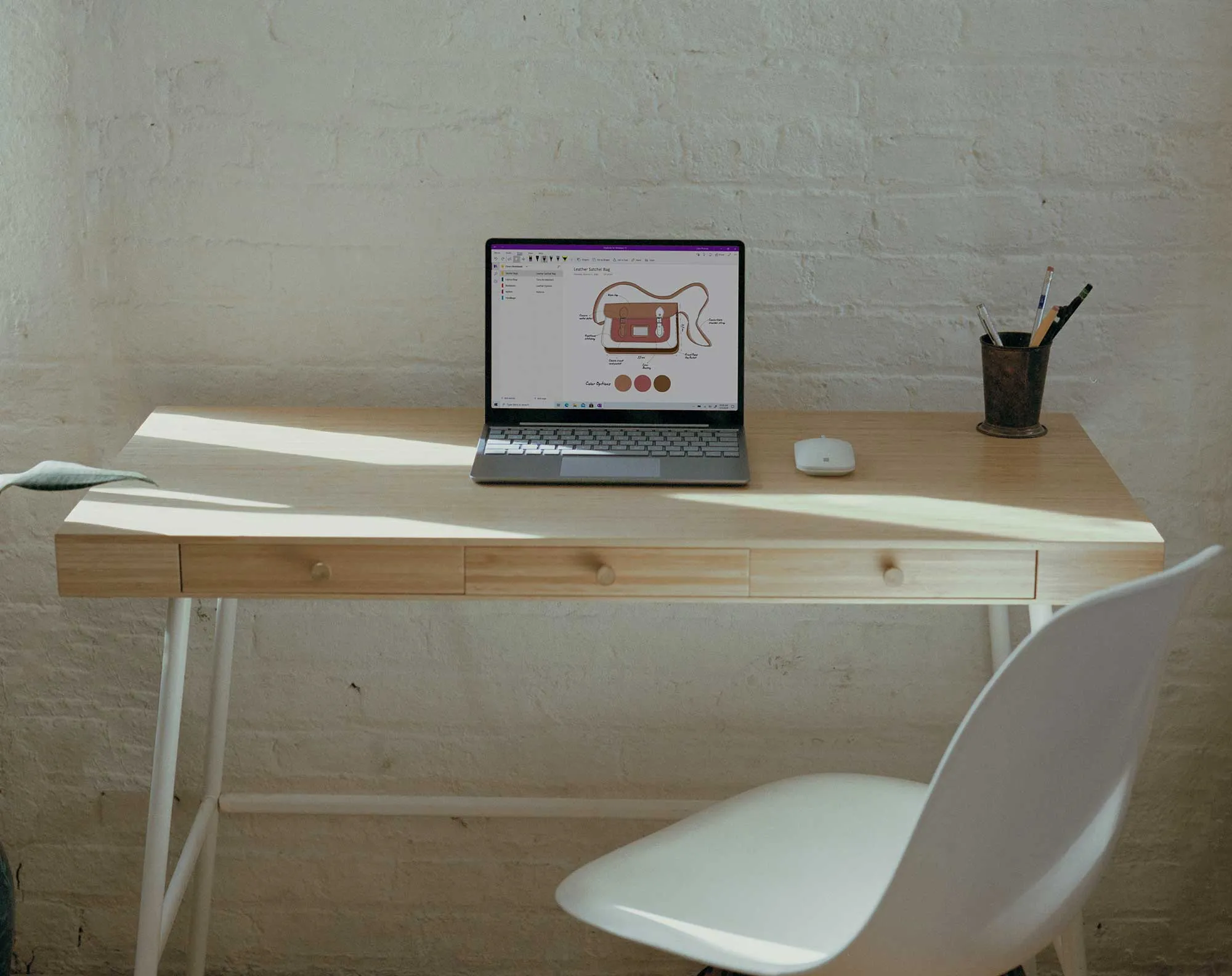 Minimalist wooden desk with a laptop displaying a leather satchel bag design, a white wireless mouse, a cup with pens, and a white chair.