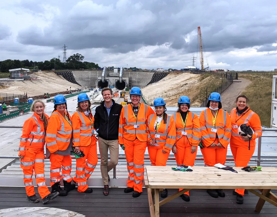 Group of nine construction workers in orange safety gear and blue helmets standing outdoors at a construction site with tunnels and machinery in the background.