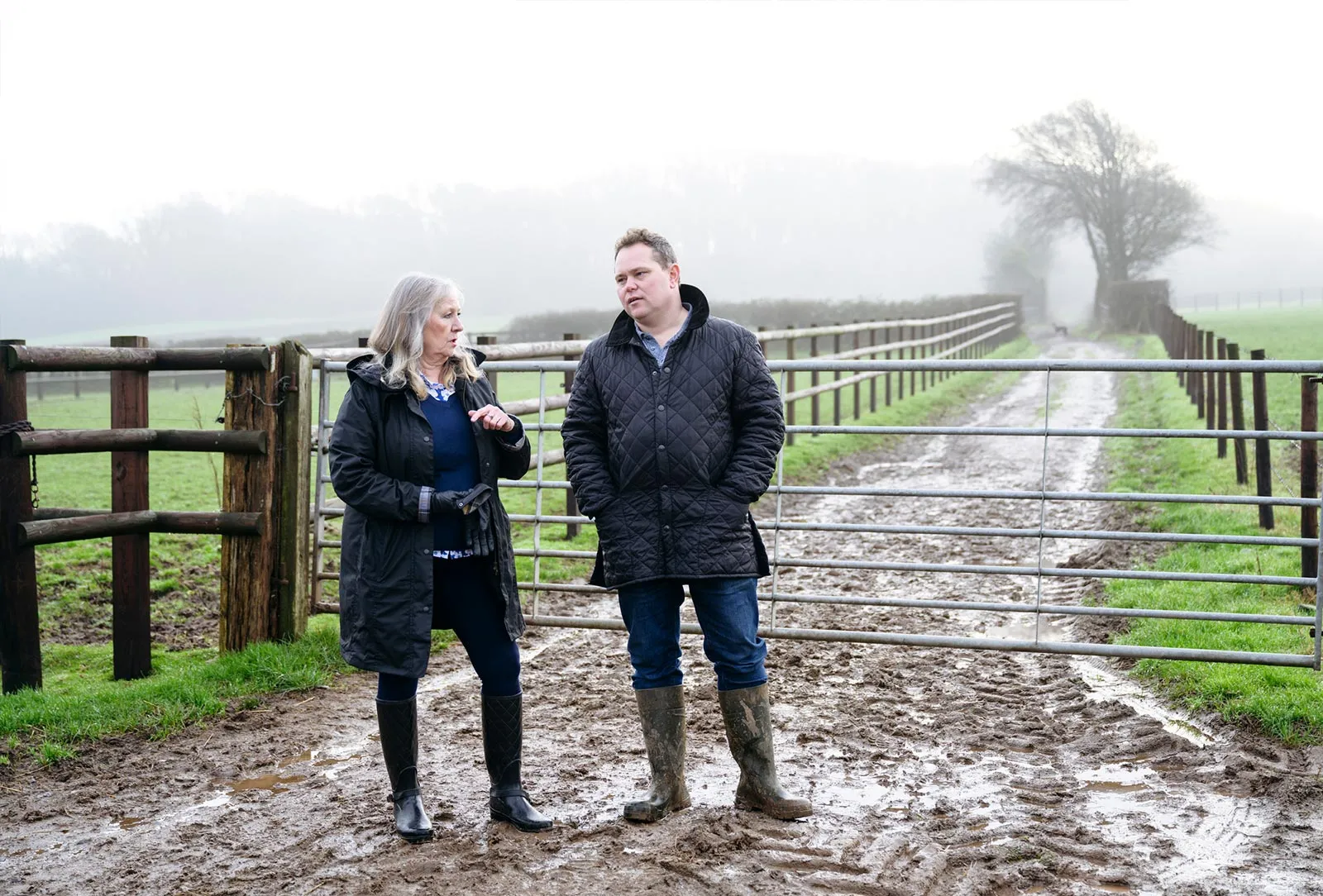 A middle-aged woman and man in raincoats and boots standing on a muddy farm path near a metal gate and wooden fence, talking.