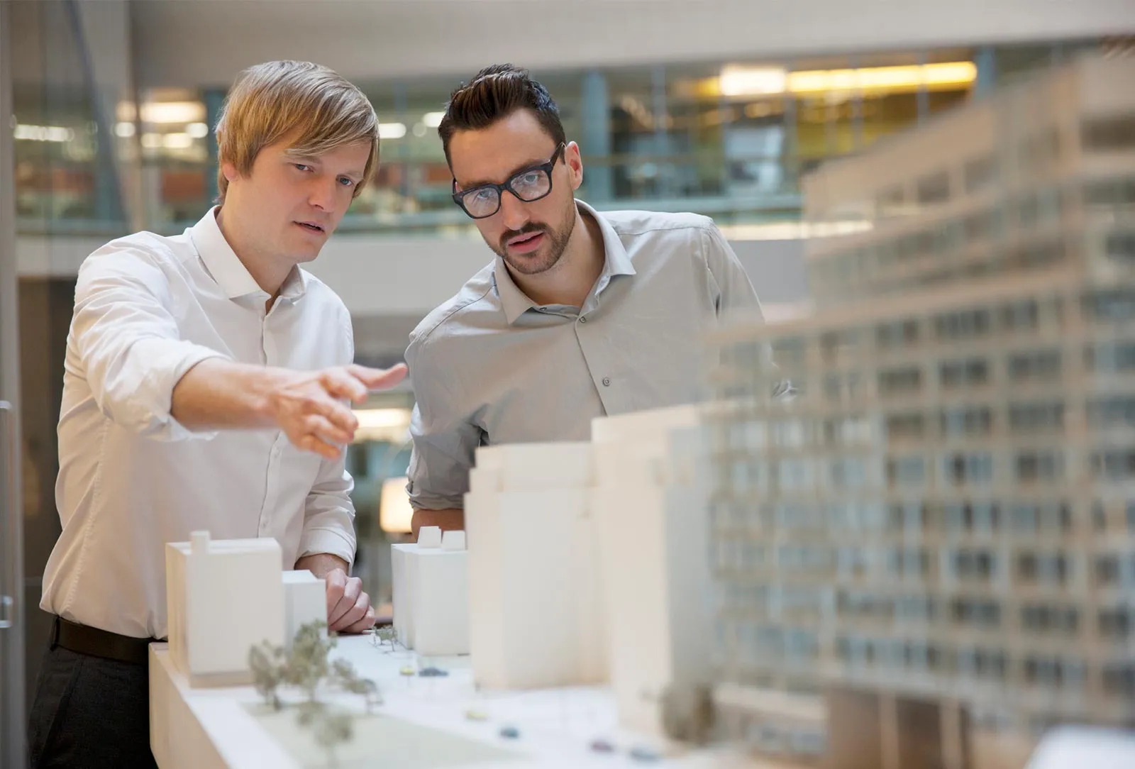 Two male architects or planners in white and light gray shirts examining a detailed architectural building model indoors.