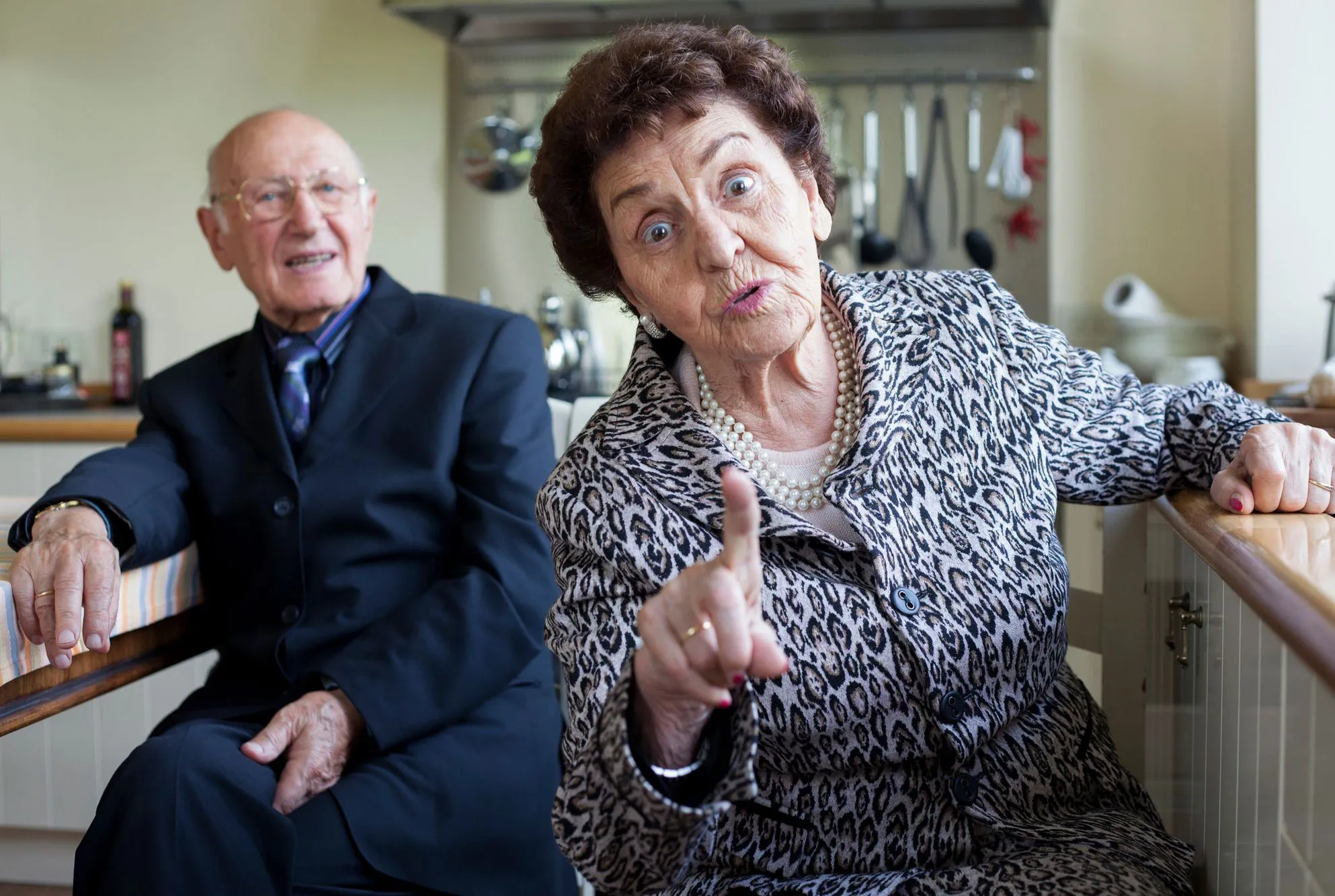 Elderly woman in patterned coat pointing her finger forward with an expressive face, with an elderly man in a suit sitting in the background.