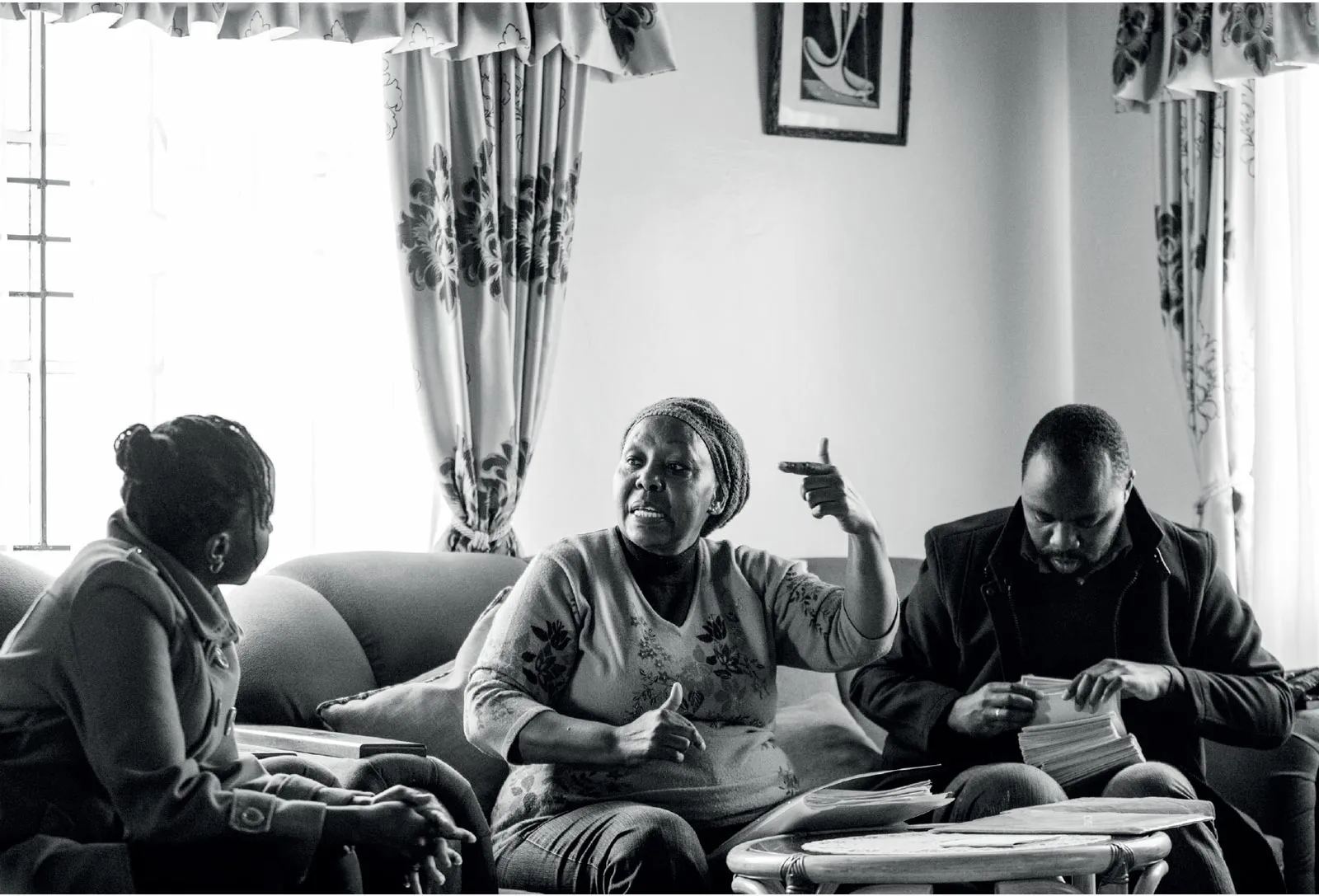 Three people sitting on a couch in a living room; a woman in the middle is speaking emphatically while gesturing with her hands, another woman listens attentively, and a man looks through papers.