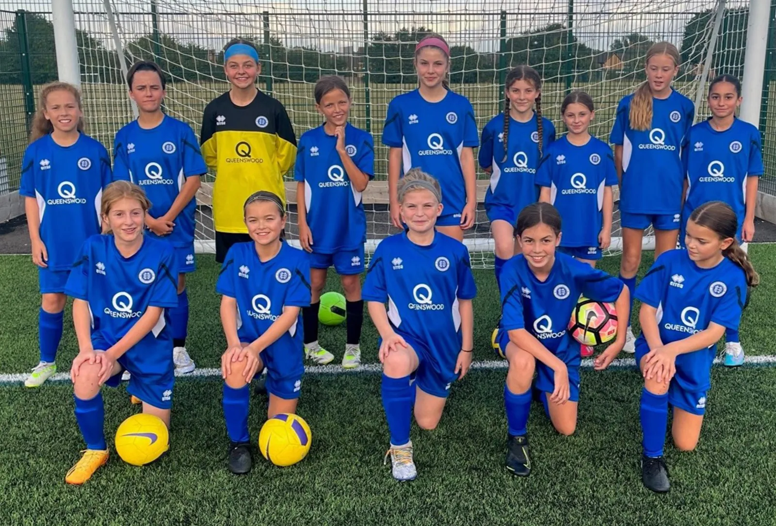 Youth girls soccer team posing on a field in blue Queenswood uniforms with four soccer balls, standing and kneeling in front of a goal.