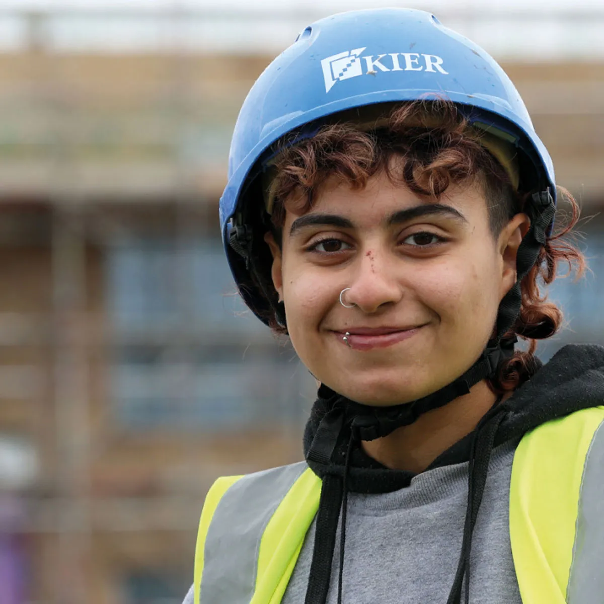 Person wearing a blue Kier safety helmet and yellow reflective vest smiling at a construction site.