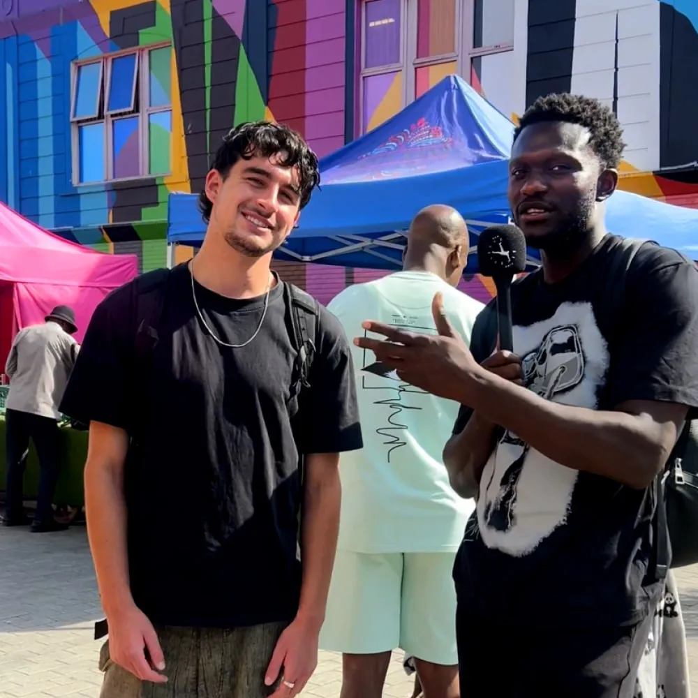 Two men standing outdoors with colorful mural and market tents in the background, one holding a microphone and gesturing.
