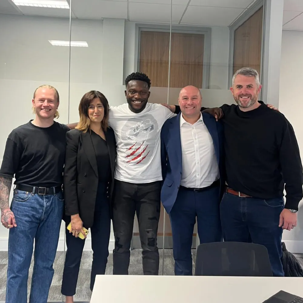 Five diverse adults standing closely in an office setting, smiling at the camera with arms around each other.