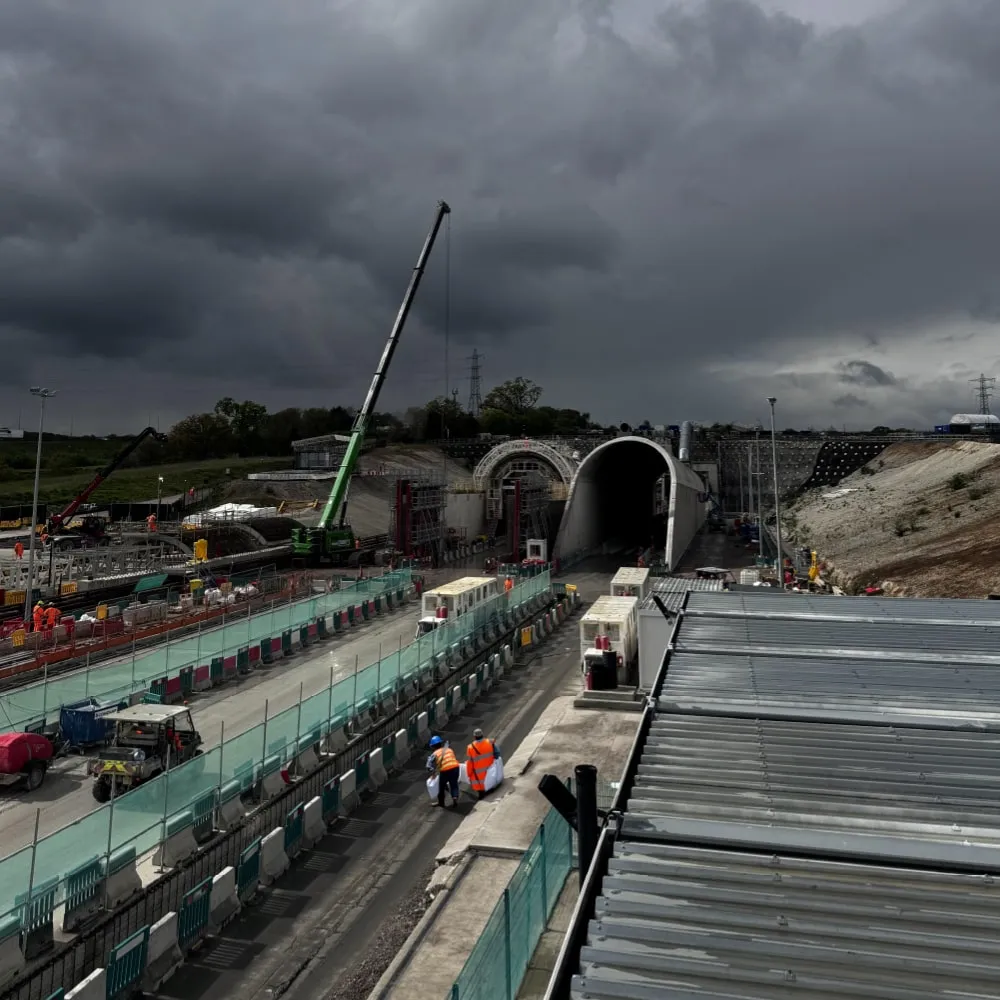 Construction site with workers in orange vests, cranes, and a large tunnel under a dark cloudy sky.