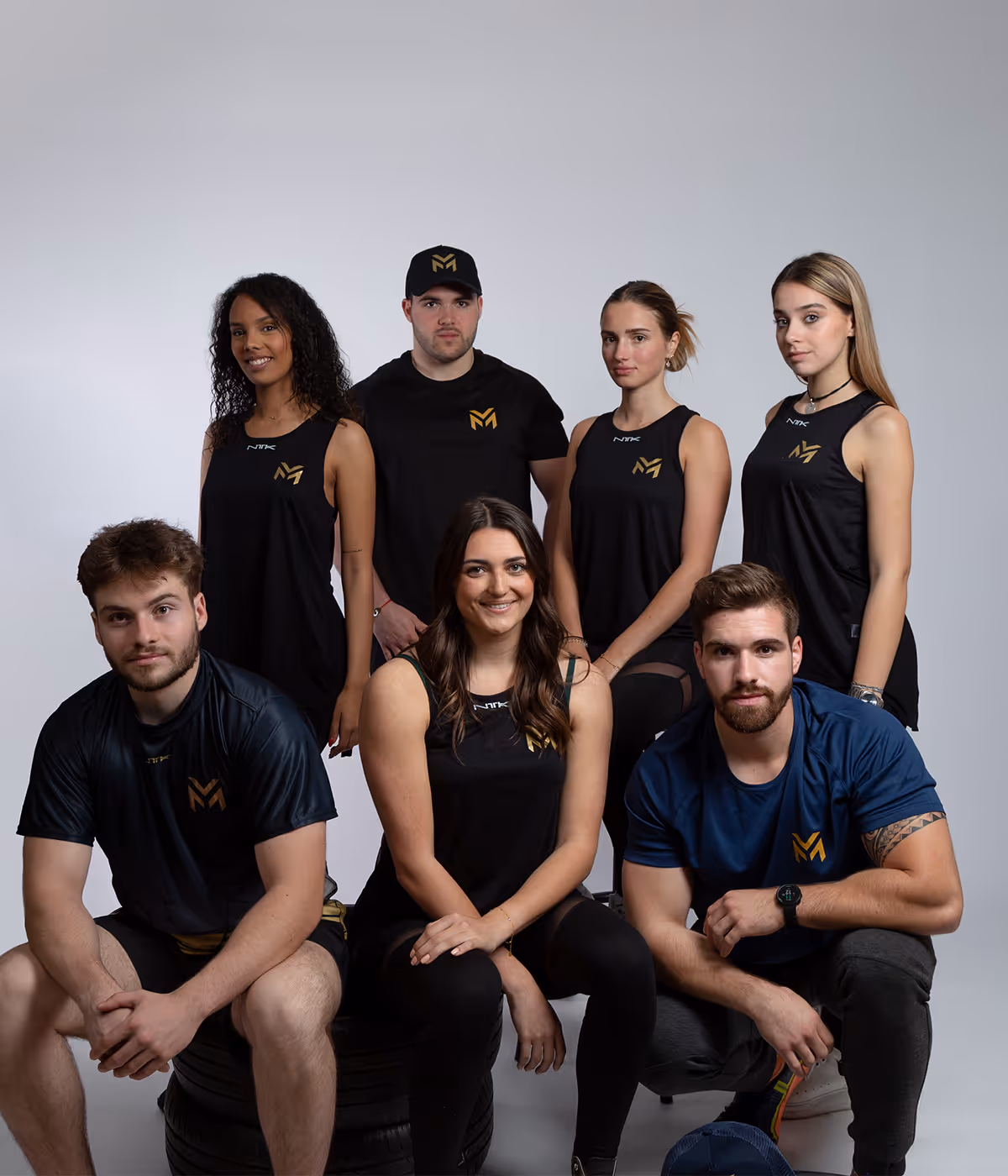 Groupe de sept jeunes adultes portant des tenues de sport noires avec logo doré, posant ensemble sur un fond gris clair.