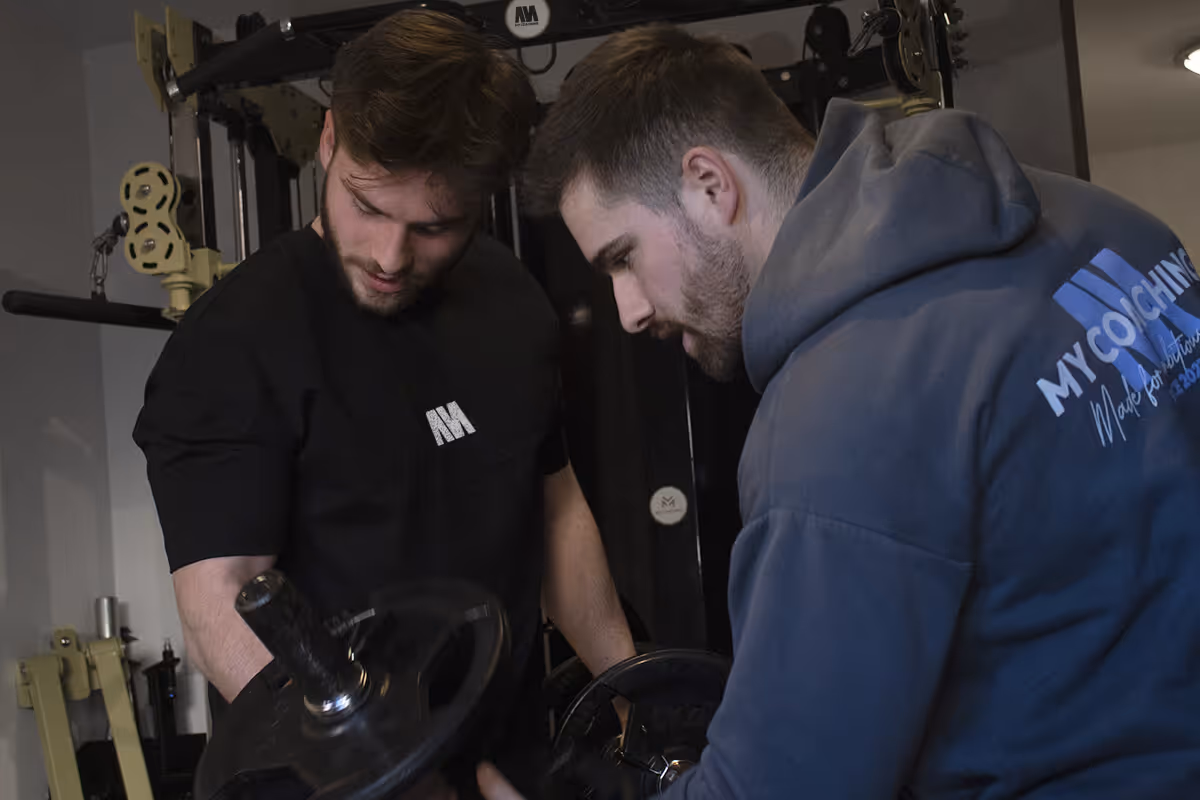 Yoann Mouly faisant un coaching en présentiel. Le client soulève une haltère dans chaque main.