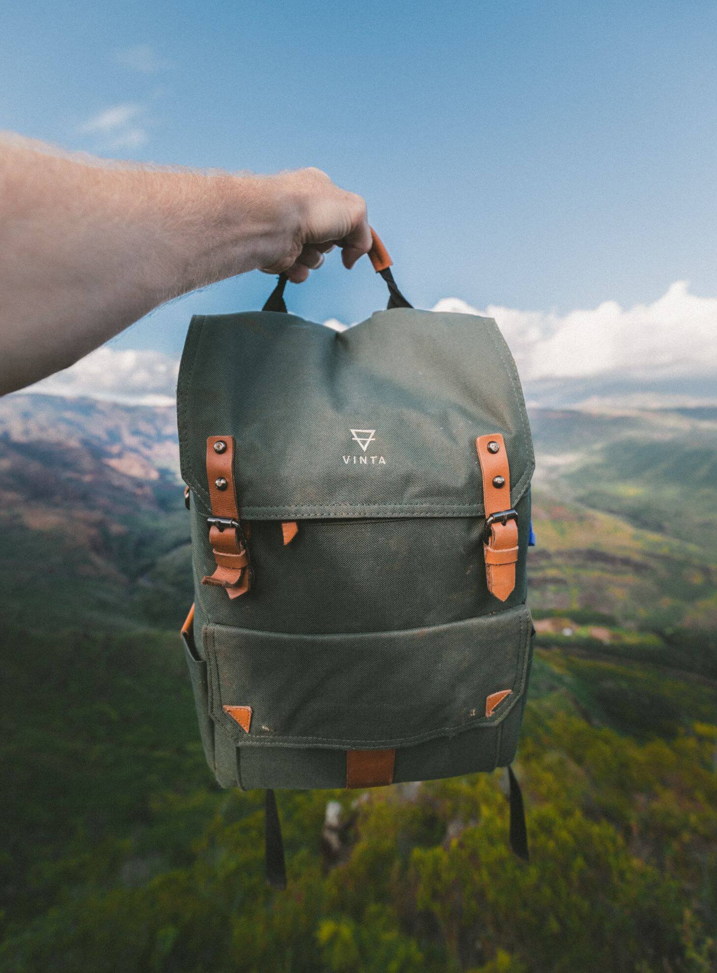 Hand holding a green Vinta backpack with leather straps against a mountain landscape background under a blue sky.