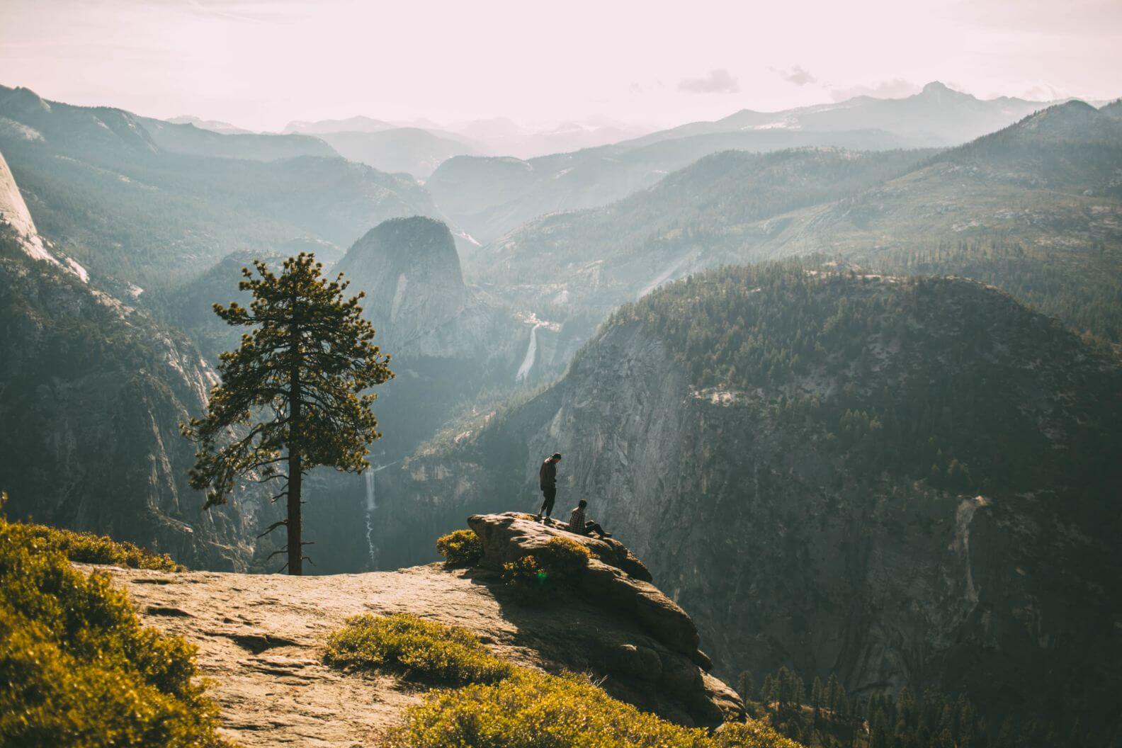 Two people on a rocky cliff overlooking a mountainous valley with waterfalls and pine trees at sunset.