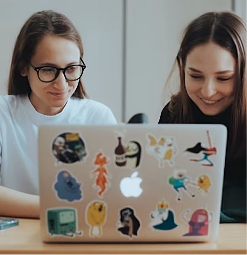 Zwei junge Frauen sitzen nebeneinander, lächeln und schauen auf einen Laptop, der mit verschiedenen bunten Aufklebern beklebt ist.