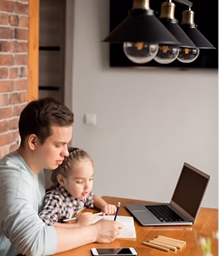 Vater hilft seiner Tochter beim Zeichnen an einem Holztisch, neben ihnen liegen ein Laptop, Stifte und ein Smartphone.