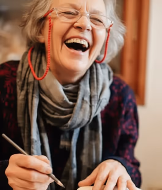 Ältere Frau mit Brille und Schal, konzentriert auf das Zeichnen in einem Skizzenbuch.