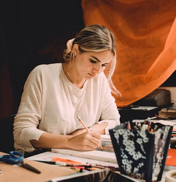 Junge Frau mit zurückgebundenen blonden Haaren, die sich auf das Zeichnen mit einem Bleistift an einem mit Malutensilien gefüllten Schreibtisch konzentriert.