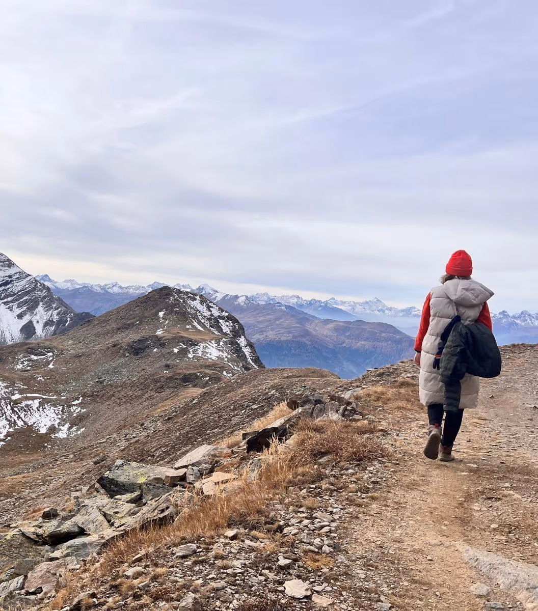 Person in a red hat and white padded vest walking on a rocky mountain trail with snow-capped peaks in the background.