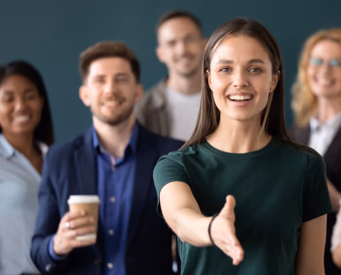 group standing looking at camera, friendly office feeling. main person is mixed race and smiling holding out her hand to shake