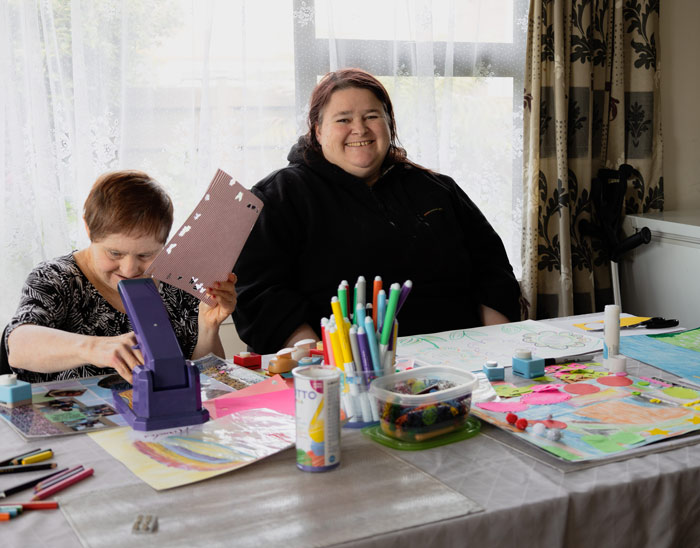 A Support Worker smiles at the camera, while helping a person they support with arts and crafts.