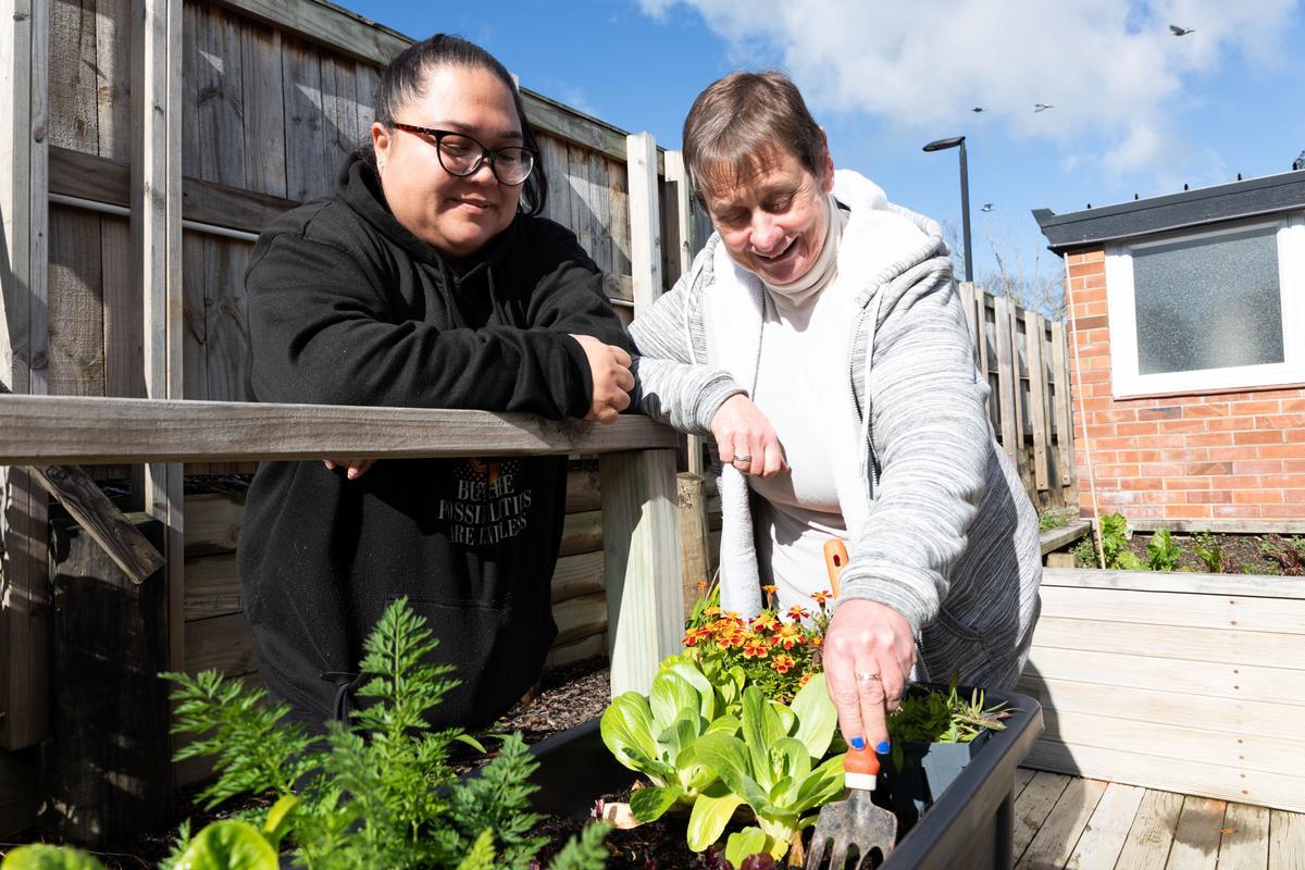 A woman tends to her vege garden while a Support Worker looks on, smiling.
